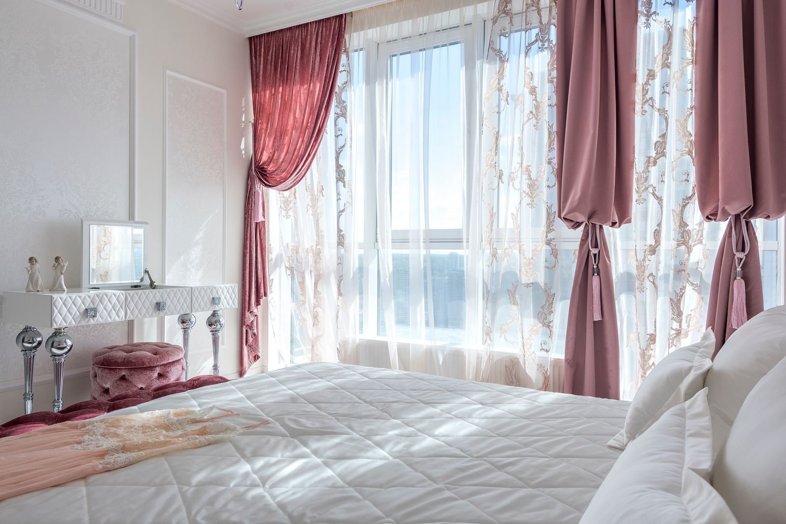 Elegant primary bedroom with floor-length rose drapery, a tufted vanity stool and warm natural light