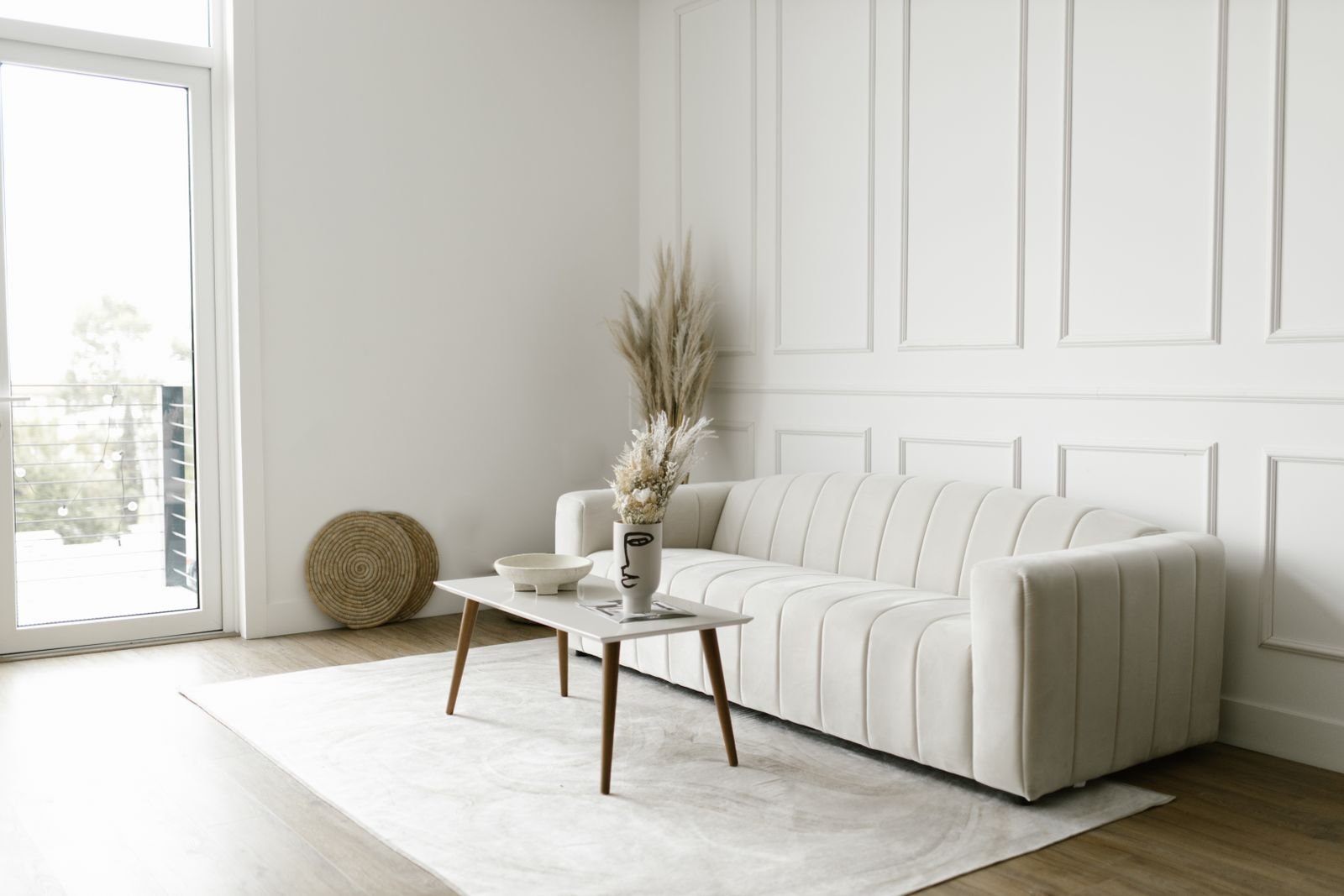 Minimalist living room with a single off-white sofa, a wood coffee table with a dried-grass vase, and a bare paneled wall