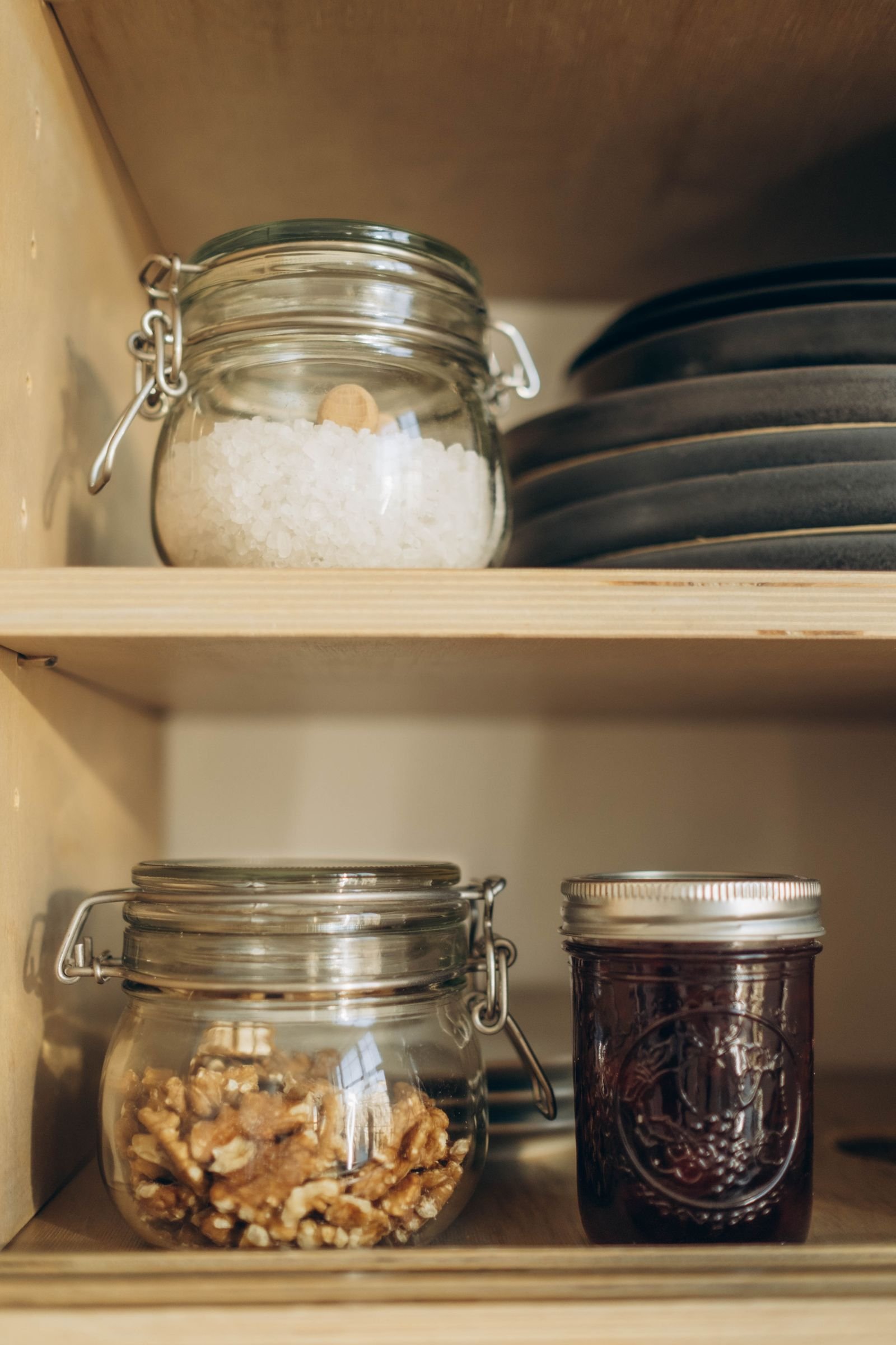 Wood pantry shelves with matching glass jars of salt and walnuts next to stacked ceramic plates